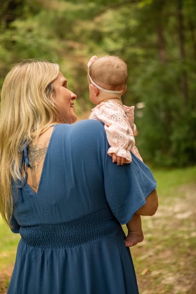 A doula carrying a baby on her shoulder along a wooded path.